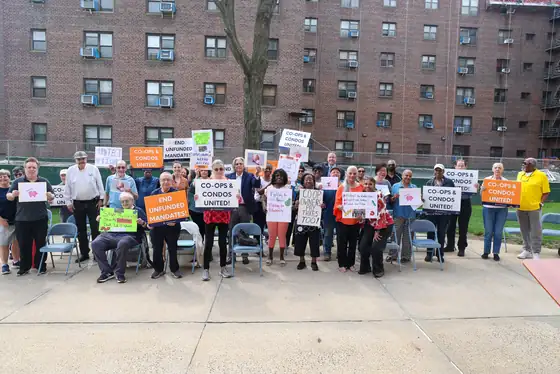 Residents standing in rows outside a large brick co-op building at Hilltop Village holding Co-ops & Condos United and End Unfunded Mandates signs
